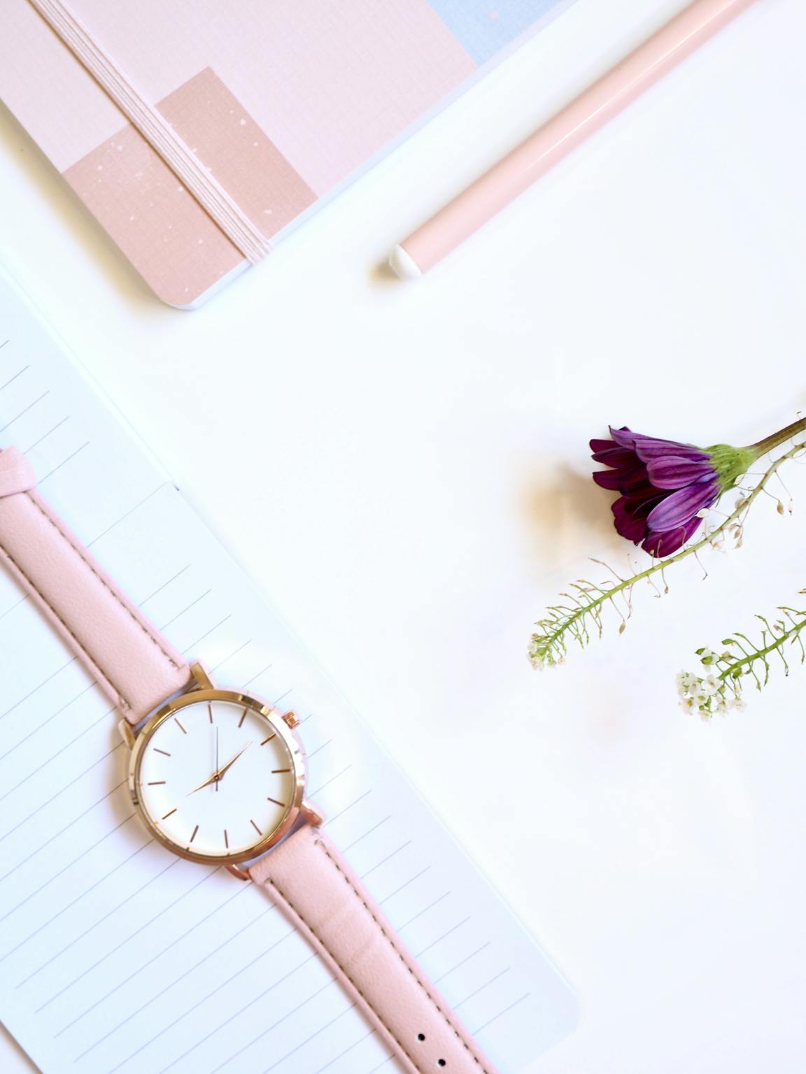 Flat lay of a minimalist pink watch with a flower and notebook on a white background.